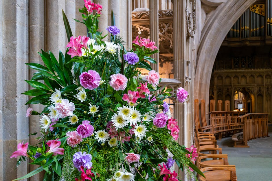 A vibrant floral arrangement featuring pink lilies, purple carnations, white daisies, and green foliage, set against the backdrop of an ornate stone interior with arched doorways and wooden pews. The 