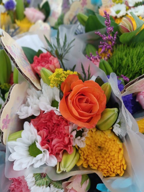 A vibrant floral arrangement featuring a prominent orange rose surrounded by white daisies, pink carnations, yellow chrysanthemums, and purple lavender sprigs, all arranged in white tissue paper with 