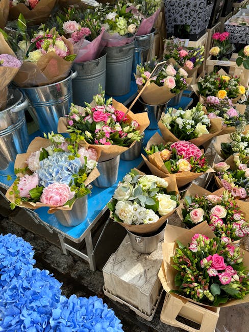 A vibrant display of freshly arranged floral bouquets featuring pastel pink roses, white roses, blue hydrangeas, and green foliage. The flowers are wrapped in brown craft paper and are presented in me