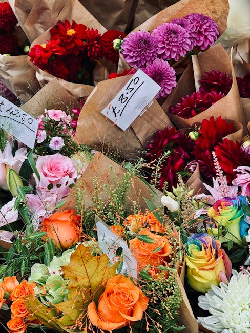 A colorful display of various fresh flowers including vibrant red dahlias, purple chrysanthemums, soft pink roses, orange roses, and multicolored spray roses arranged in small brown paper wraps. The f